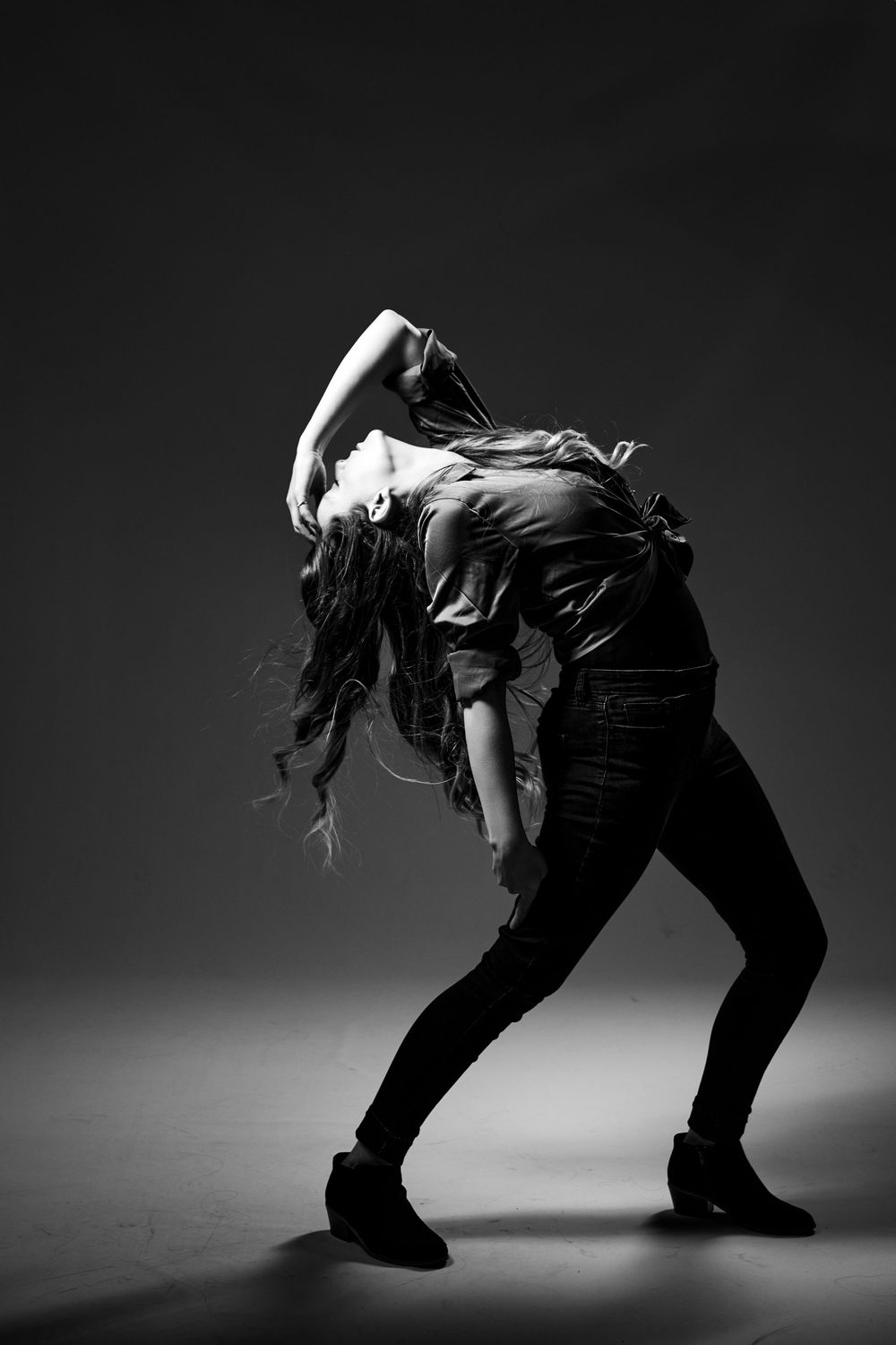 Dancer in black outfit arches backwards in dramatic pose against dark background in black and white photo.