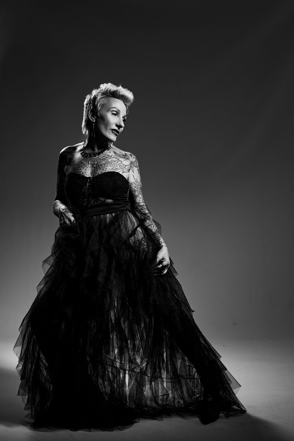 A dramatic black and white portrait of a figure in an elegant black tulle evening gown against a dark background.