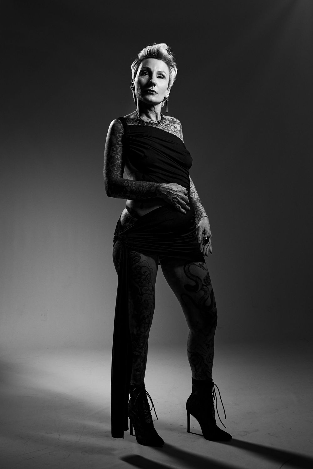 Black and white dramatic studio portrait of a person in stilettos and dark outfit against gradient background.