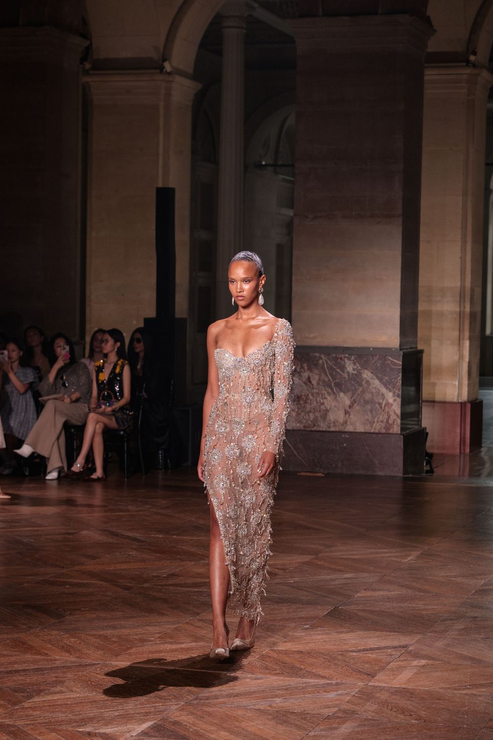 Six sequential fashion runway photos showing a shimmering silver beaded evening gown in motion on a stone floor.