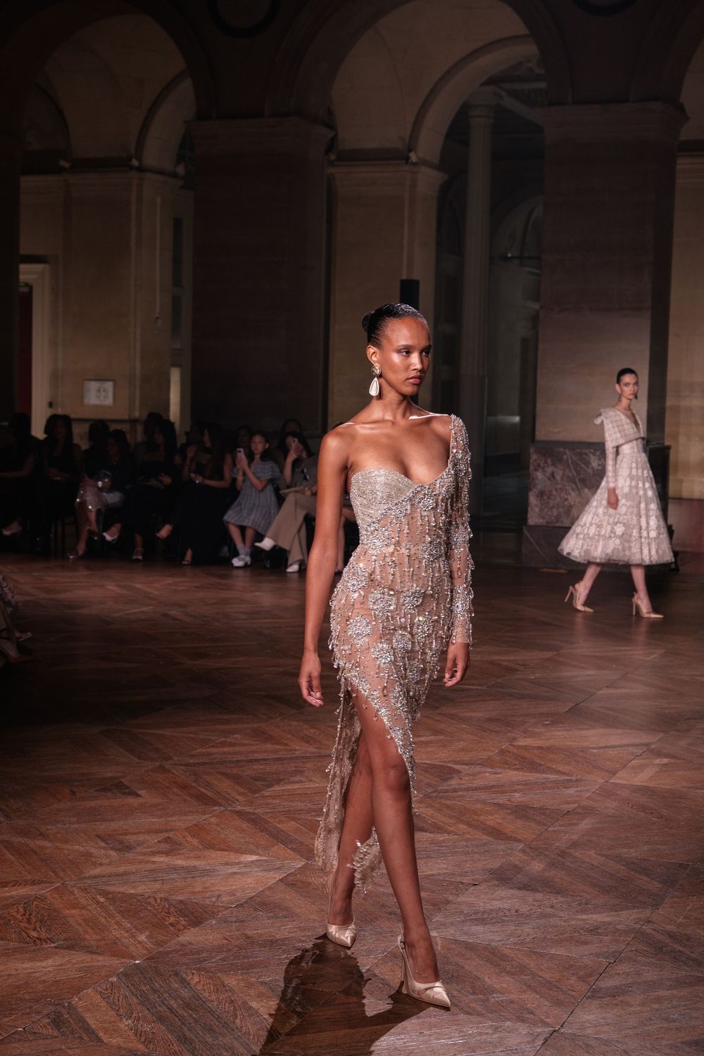 Model walks runway in glittering sheer crystal dress during haute couture fashion show in grand architectural space.