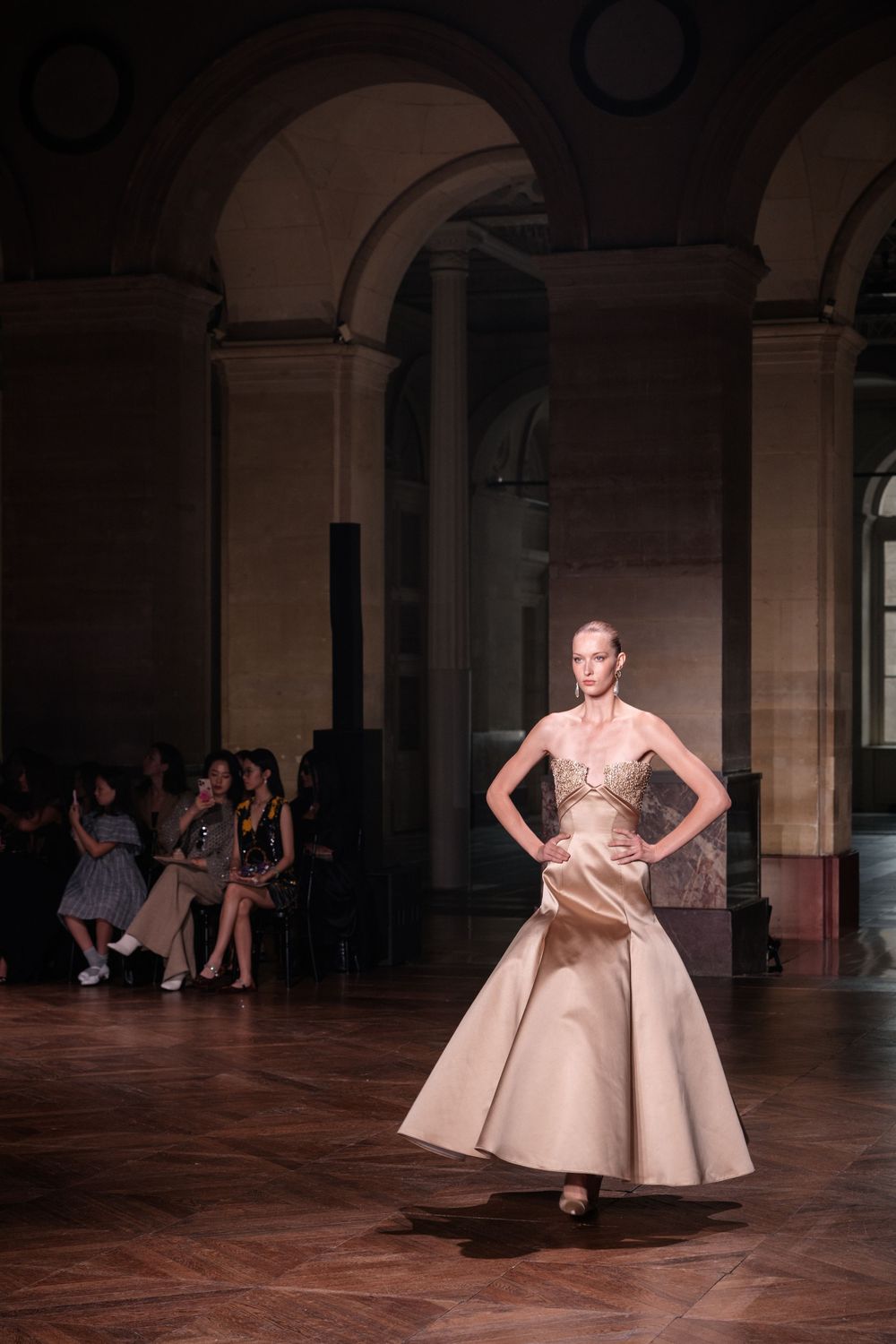A dancer in a beige dress performs an elegant twirling movement on a polished wooden floor in a grand hall with arches.