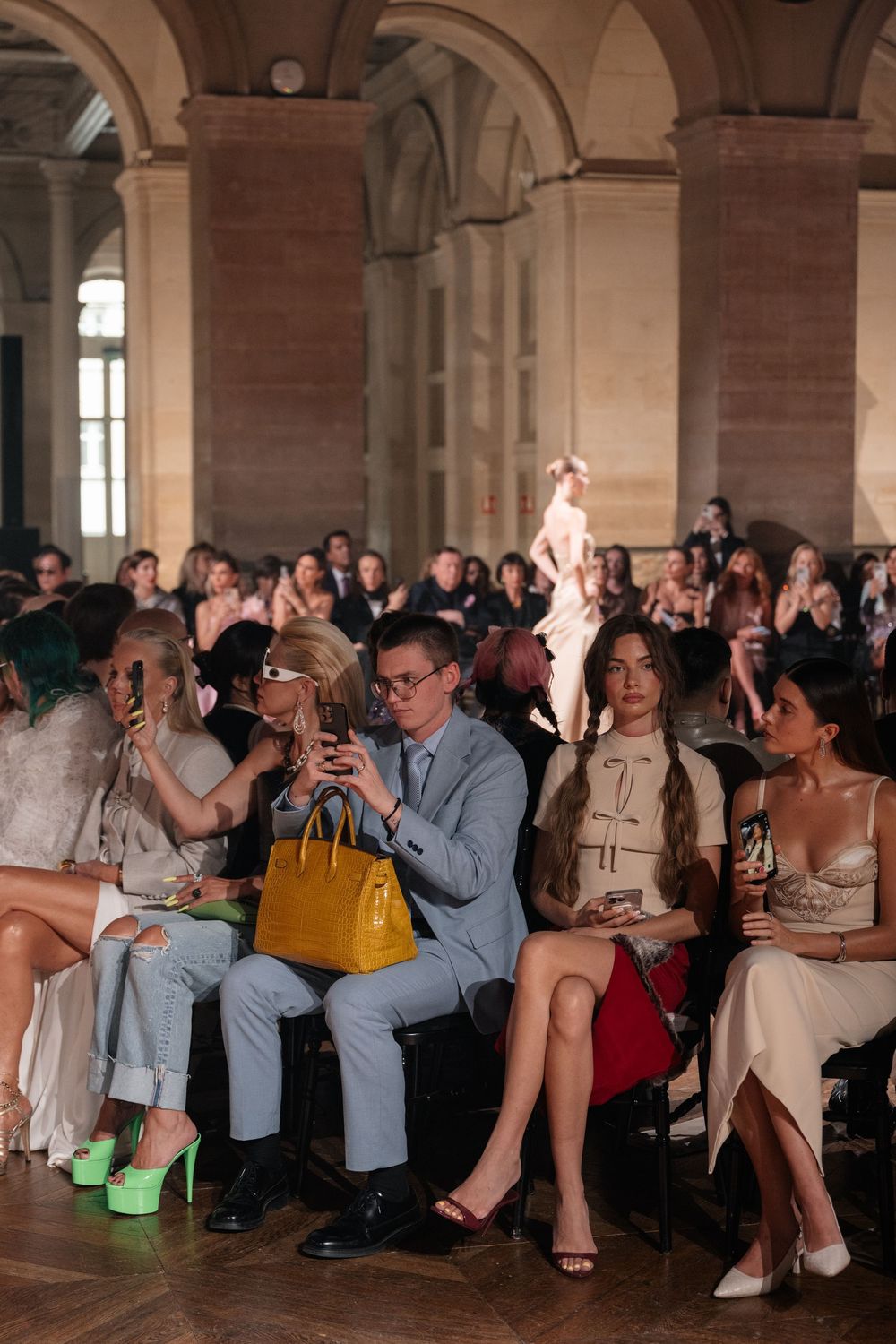 Fashion show audience members take photos and watch attentively in an elegant columned venue.