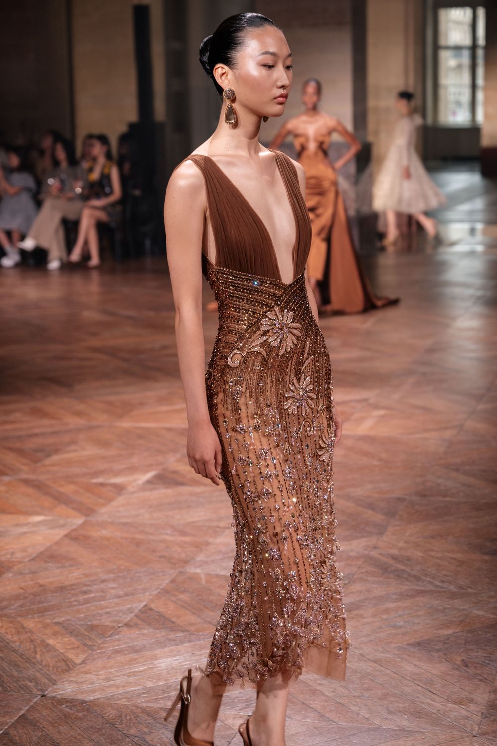 Model walks runway in elegant brown velvet and sequined midi dress with plunging neckline at fashion show.