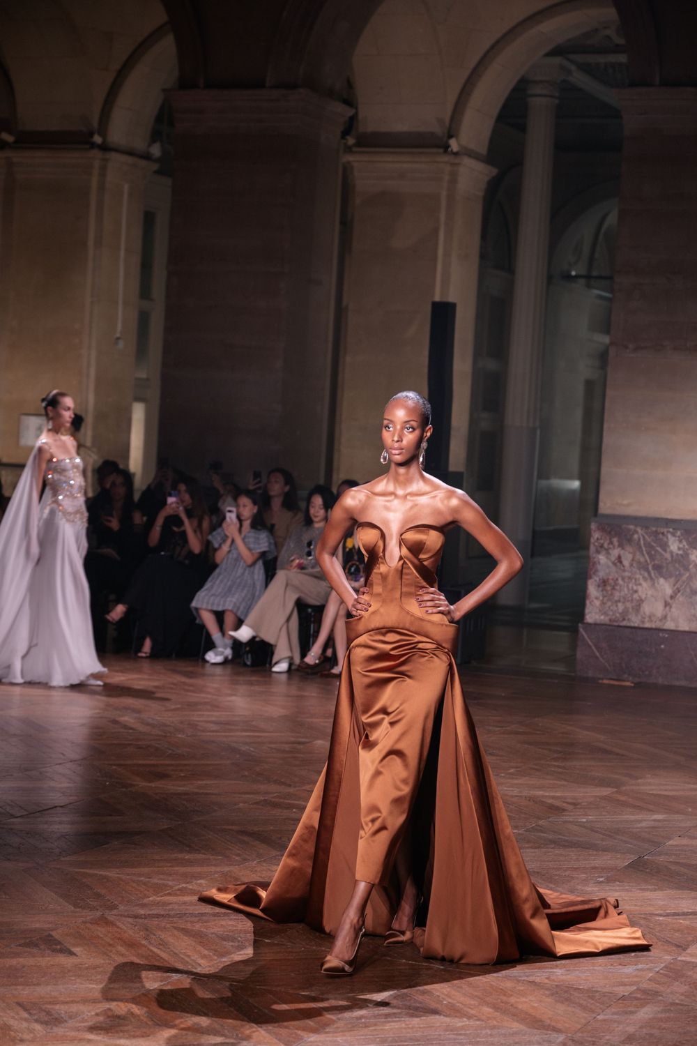 A fashion model walks the runway in a flowing copper-colored satin gown at an elegant indoor venue.