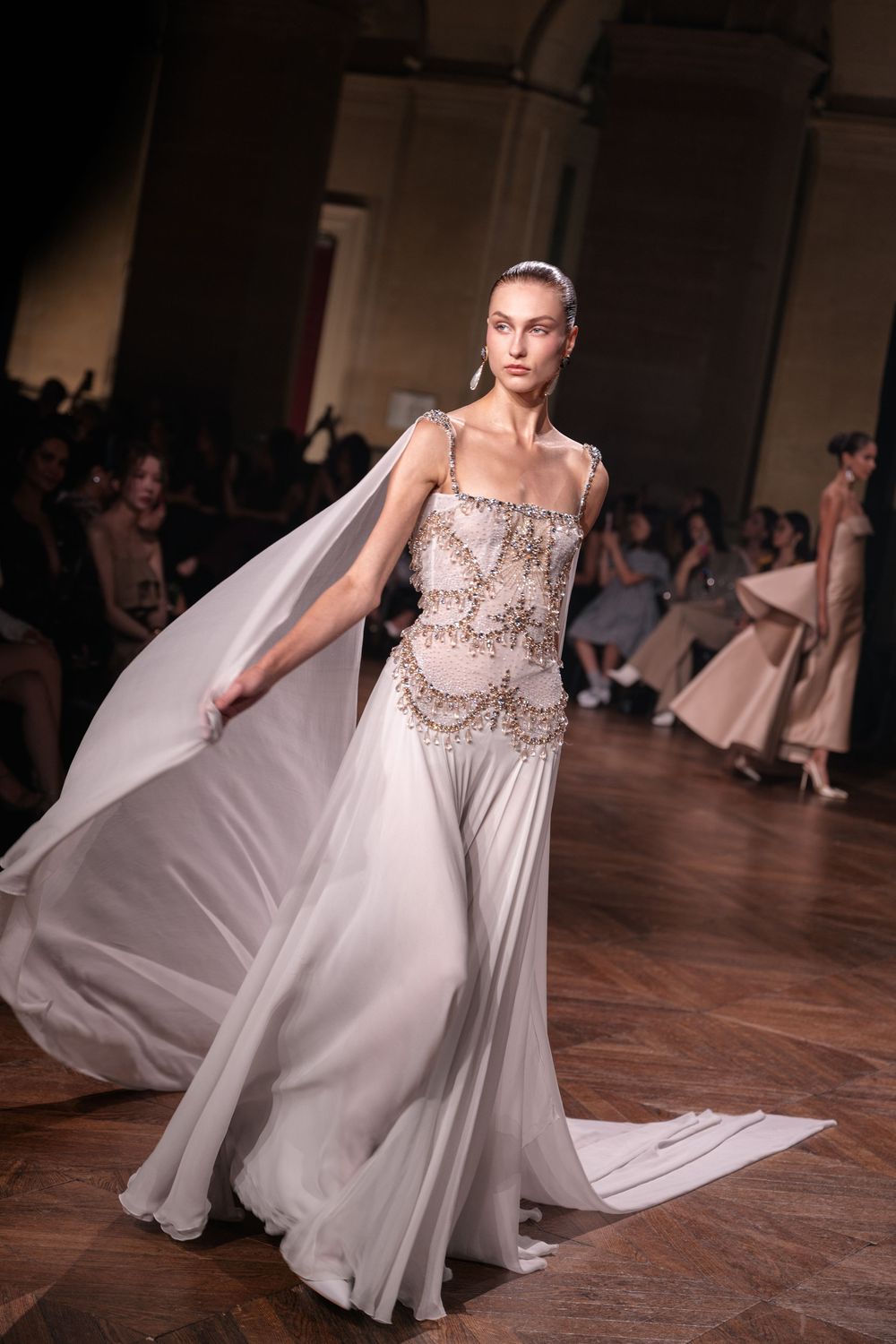 A flowing white wedding gown with beaded bodice and cape flows dramatically on runway during fashion show.