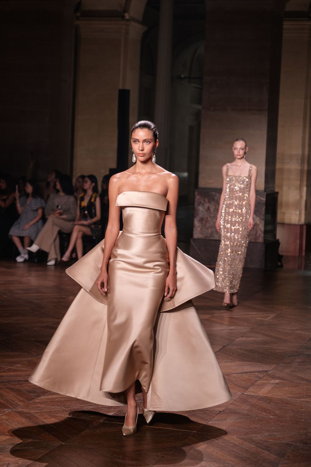 Models walk runway in elegant champagne-colored evening gowns during high fashion show on wooden parquet floor.