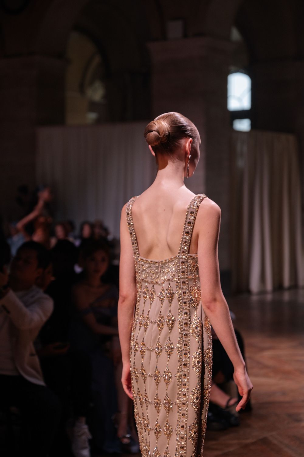 Model walks runway in beaded champagne evening gown with detailed back at luxury fashion show.