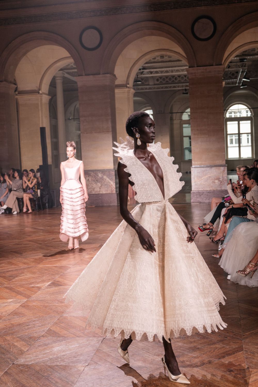 Dramatic white ball gowns with intricate detailing float down a historic arched hallway during a fashion show.
