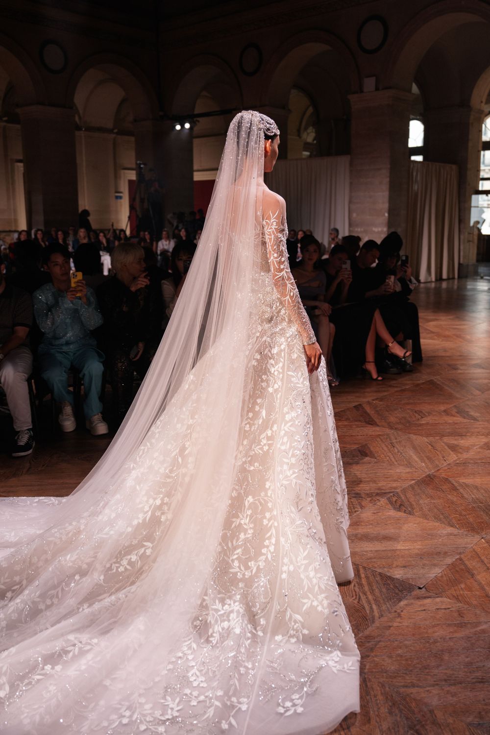 A ethereal white wedding gown with sparkling details and flowing veil trails across a grand ballroom floor.