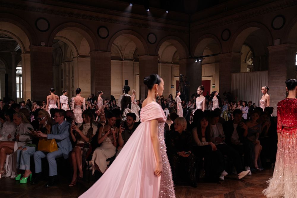 Pink flowing evening gown with dramatic cape on display at elegant indoor fashion show venue.