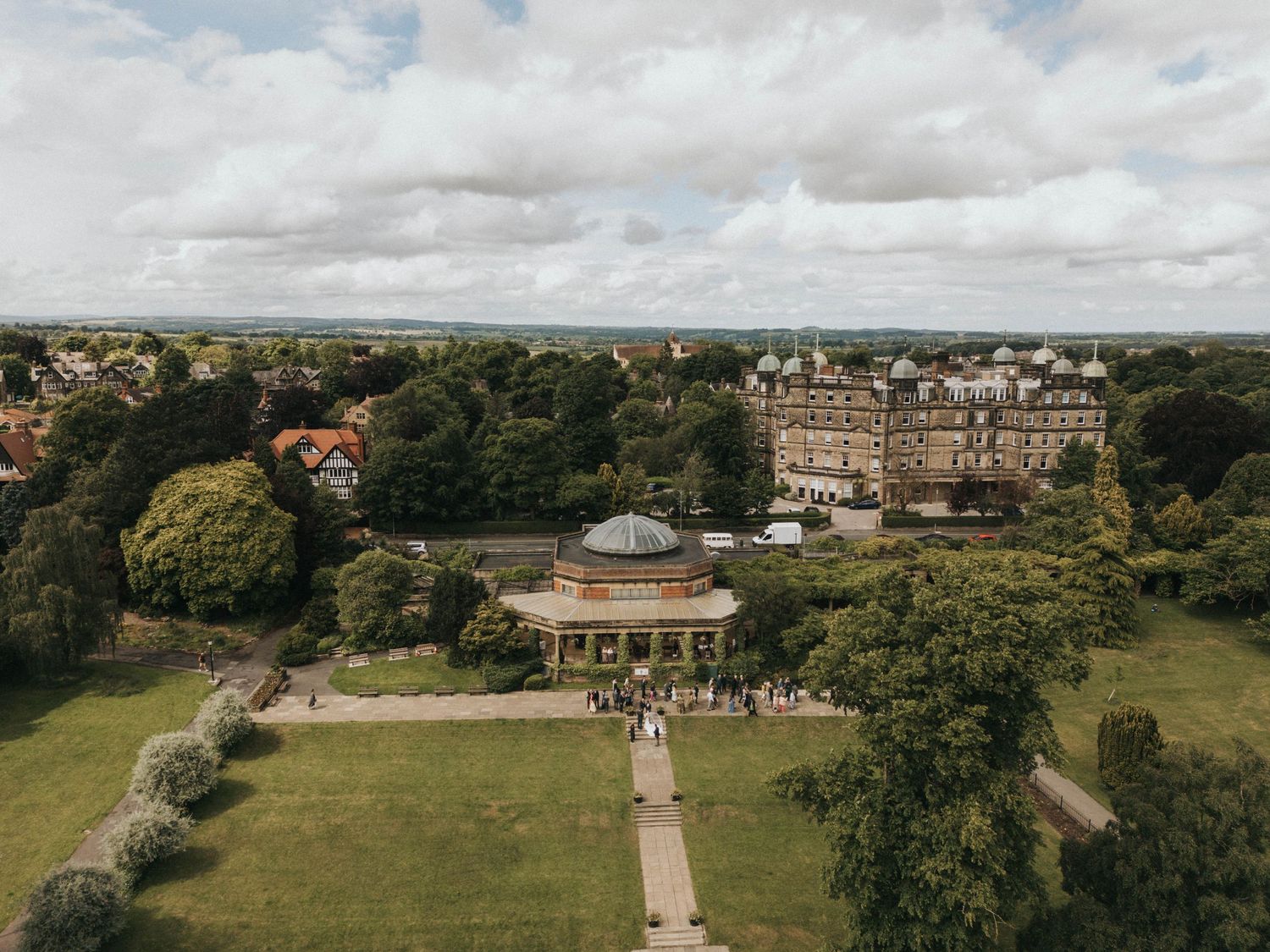 A scenic panoramic view of a historic estate with manicured gardens, stone buildings and tree-lined pathways on a cloudy day.