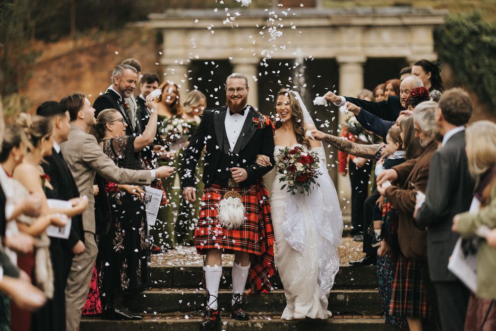 Wedding guests shower a newly married couple in traditional Scottish attire with confetti on stone steps.