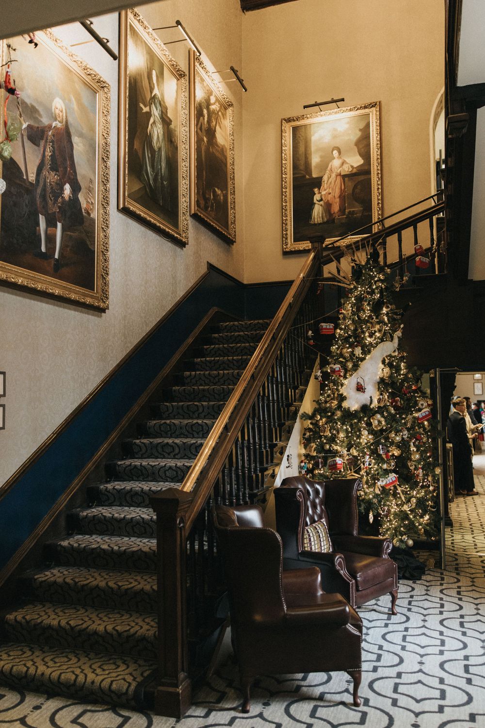 Elegant staircase with ornate portraits on wall, decorated Christmas tree, and patterned floor in historic mansion interior.