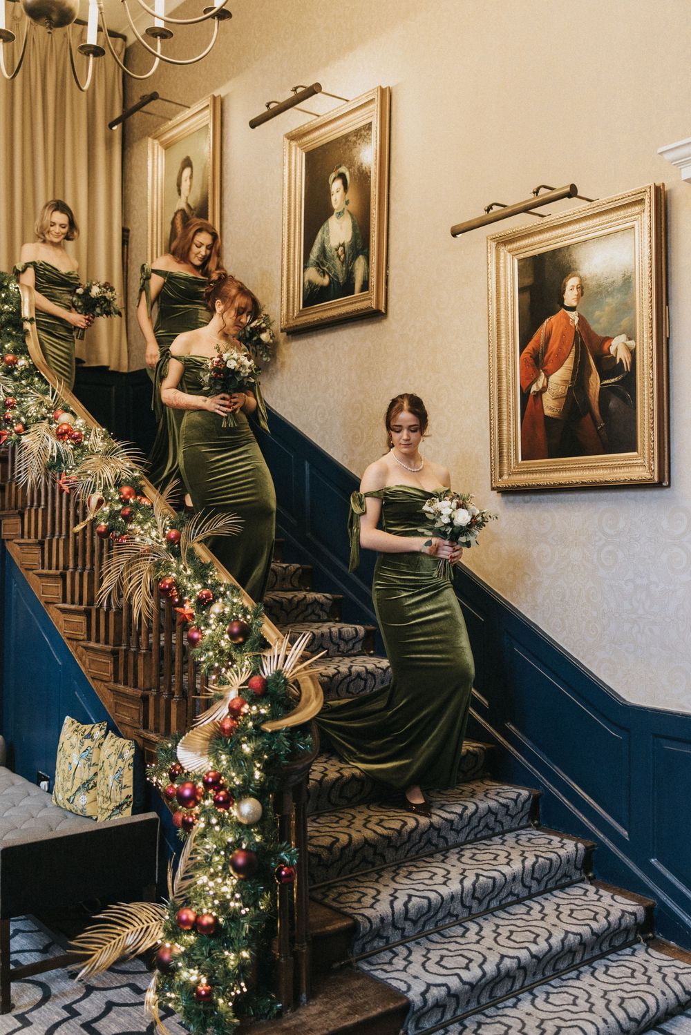 Bridesmaids in emerald green dresses pose on an elegant staircase decorated with floral garlands and historic portraits.