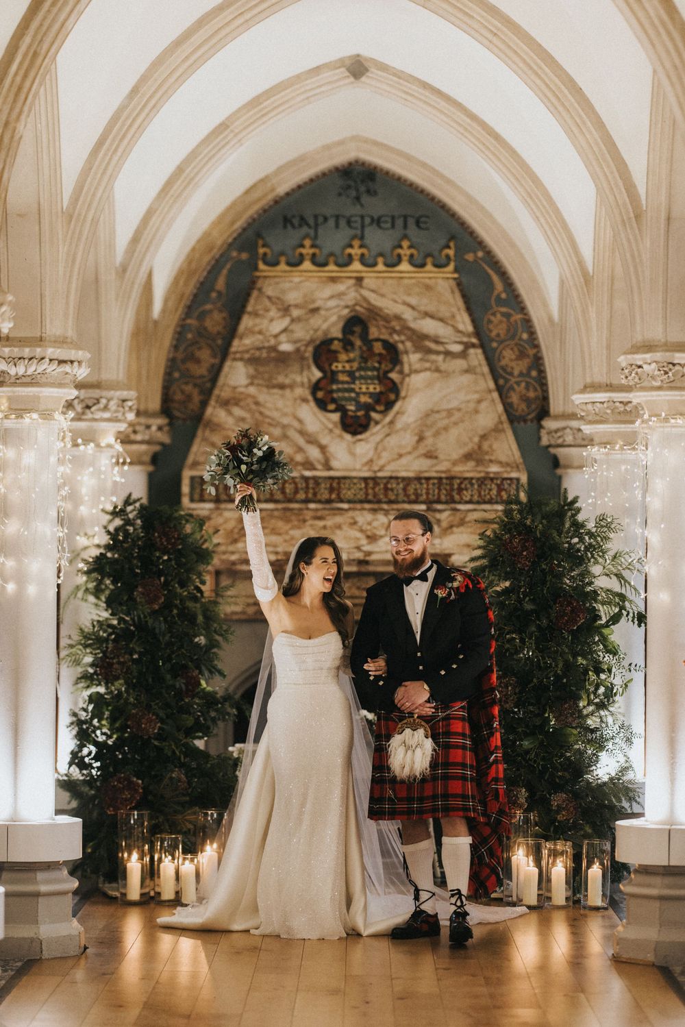 Wedding couple celebrates under ornate stone arches in a cathedral with festive greenery and lantern decorations.