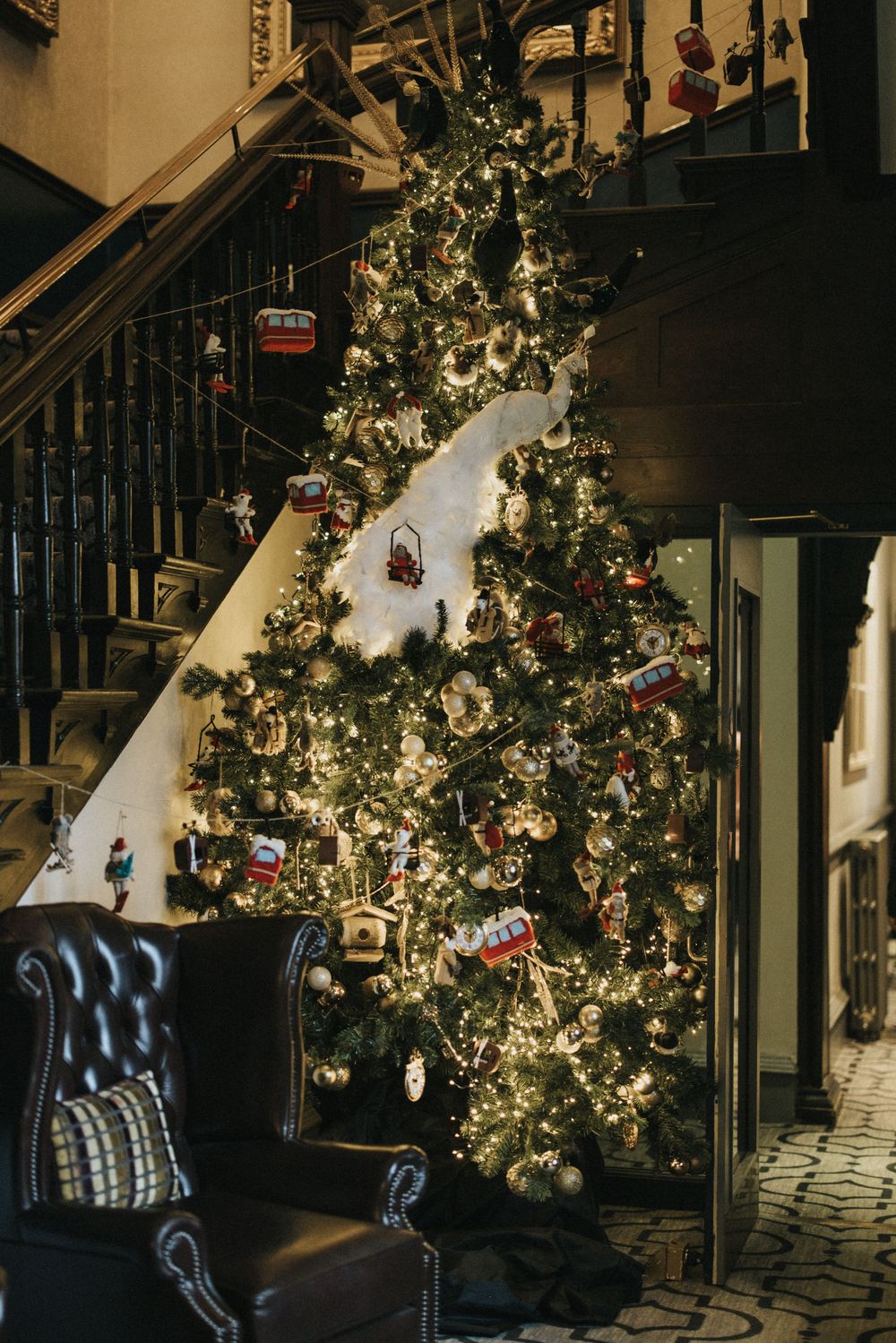 A beautifully decorated Christmas tree stands tall beside a dark staircase and leather armchair in a cozy home interior.