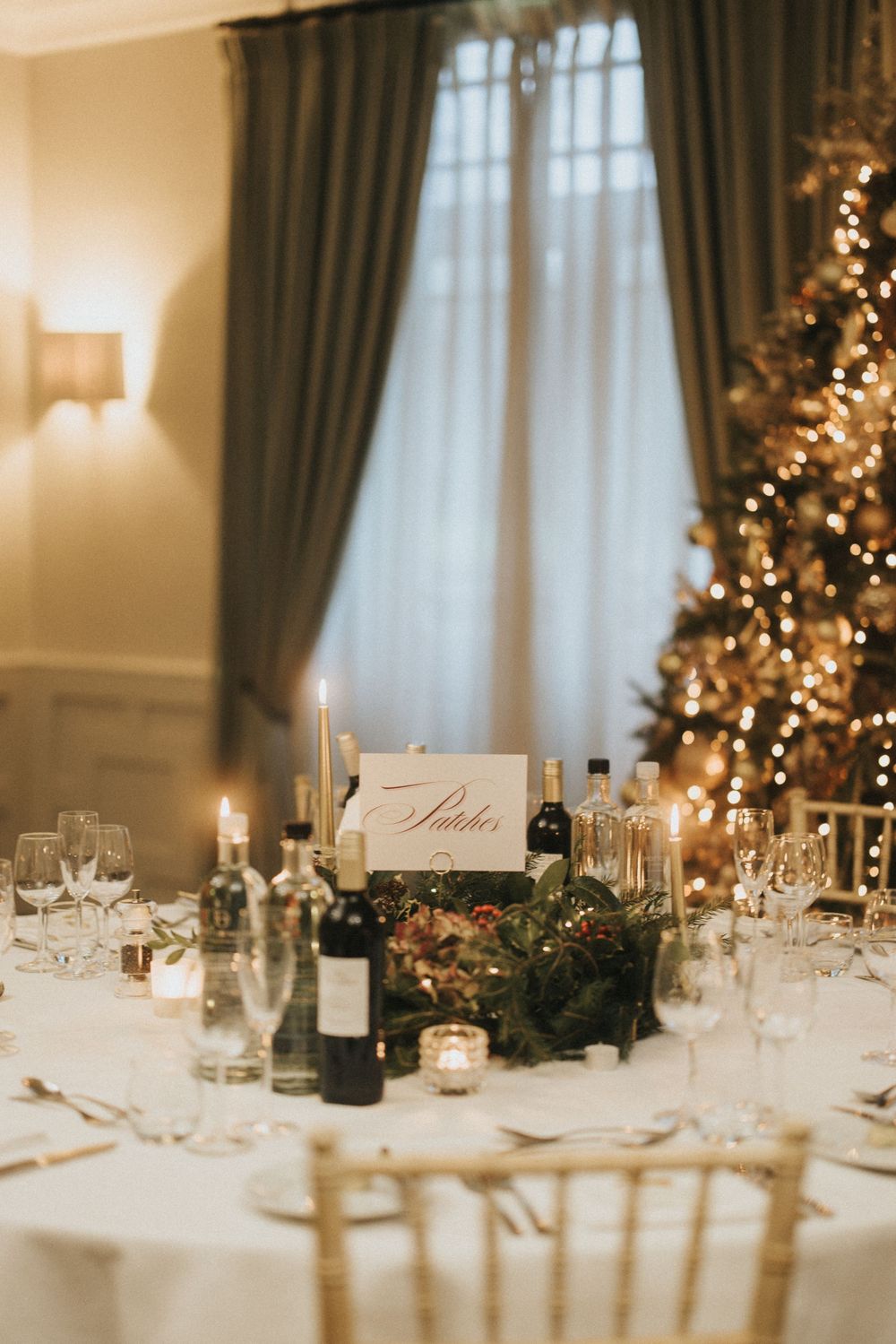 Elegant winter wedding reception table setting with white linens, candles, and Christmas tree decor in the background.