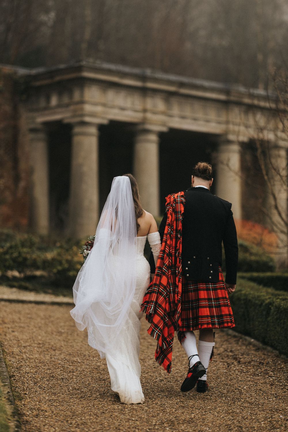 Newlywed couple walking on gravel path with groom in traditional Scottish kilt and bride in flowing white gown with veil.