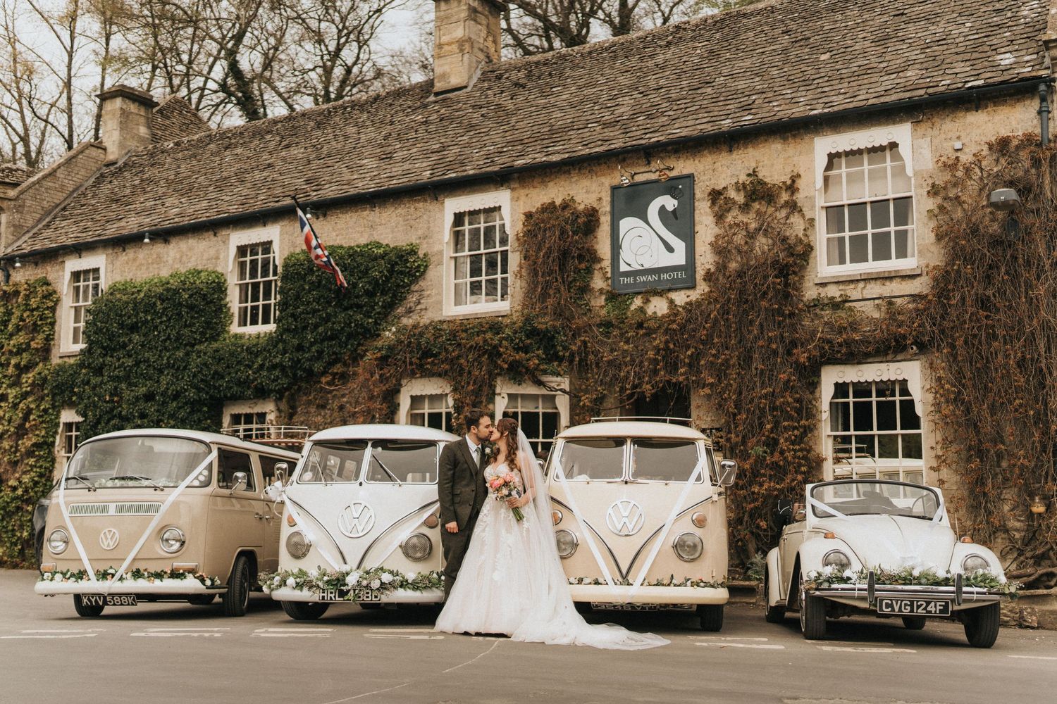 Three vintage Volkswagen buses and a classic car parked in front of an ivy-covered stone cottage with wedding attire.