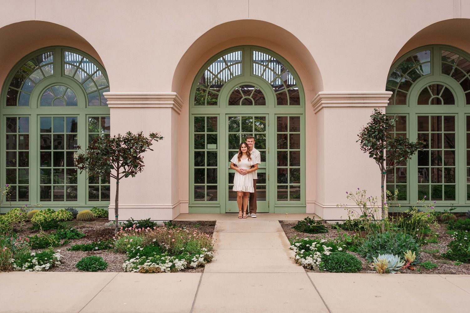Someone stands between three elegant arched French doors surrounded by a manicured garden and pink stucco facade.