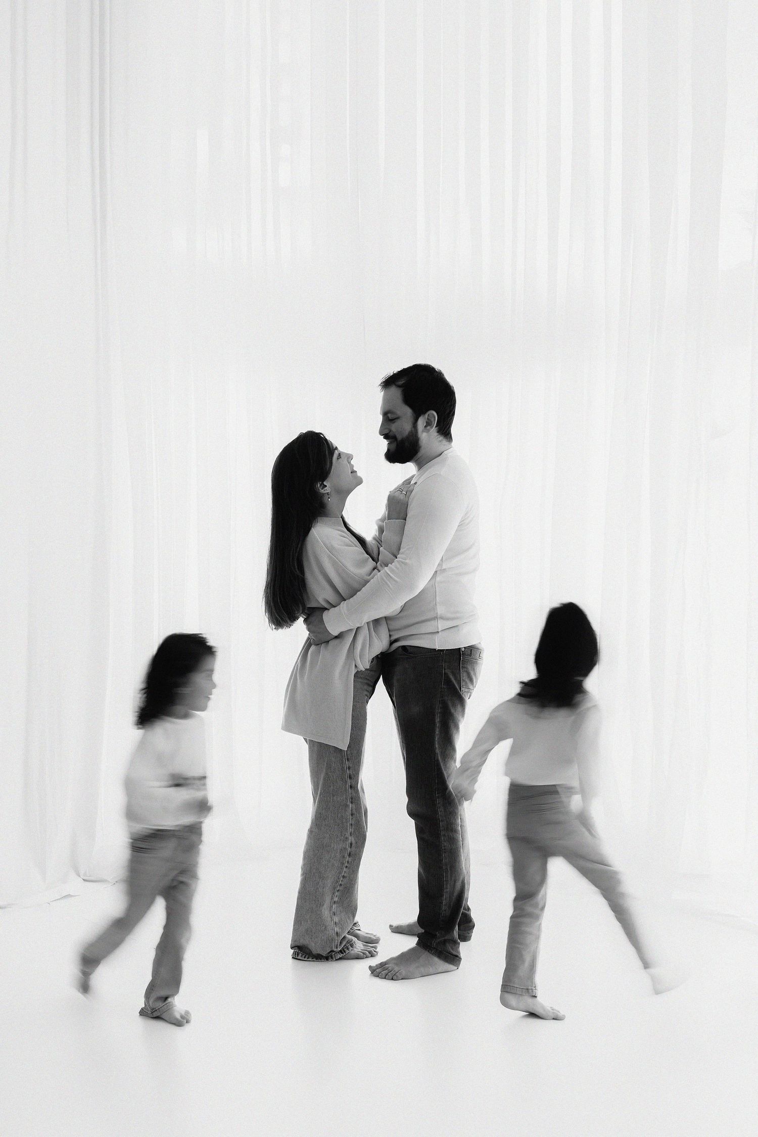 Black and white family portrait showing grown-ups embracing while blurred children run past them against white backdrop.