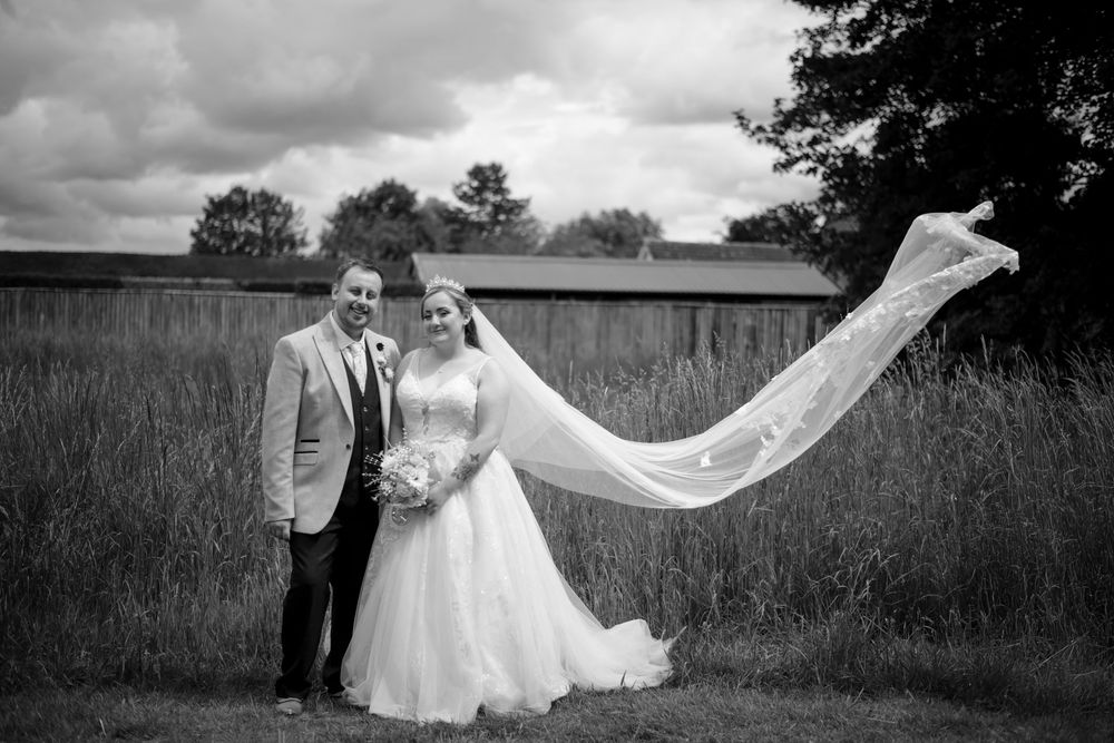 A dramatic wedding photo in black and white shows a bride's veil flowing in the wind against a rustic countryside backdrop.