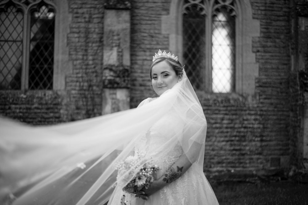 A bride in a flowing veil poses outside a historic brick church with gothic windows in black and white.