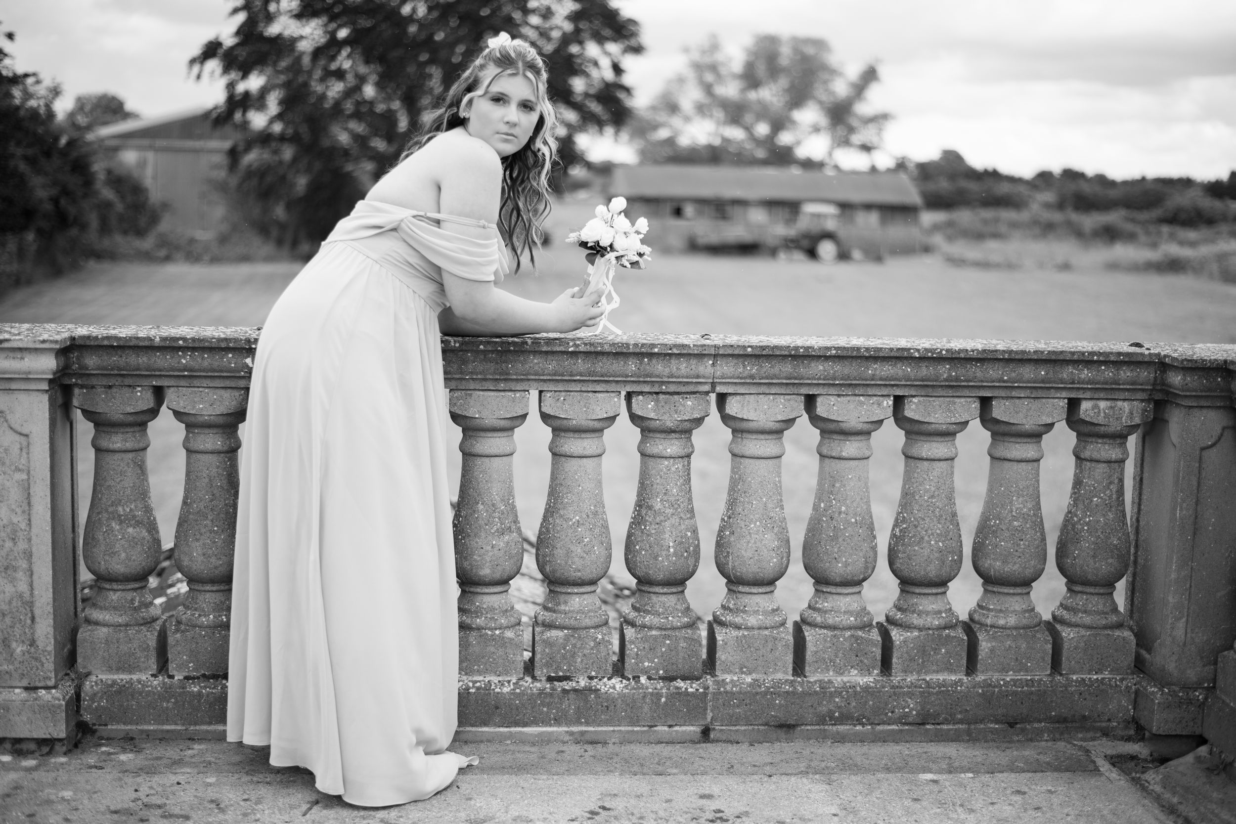 A black and white photograph showing someone in a flowing white dress leaning over an ornate stone balustrade.