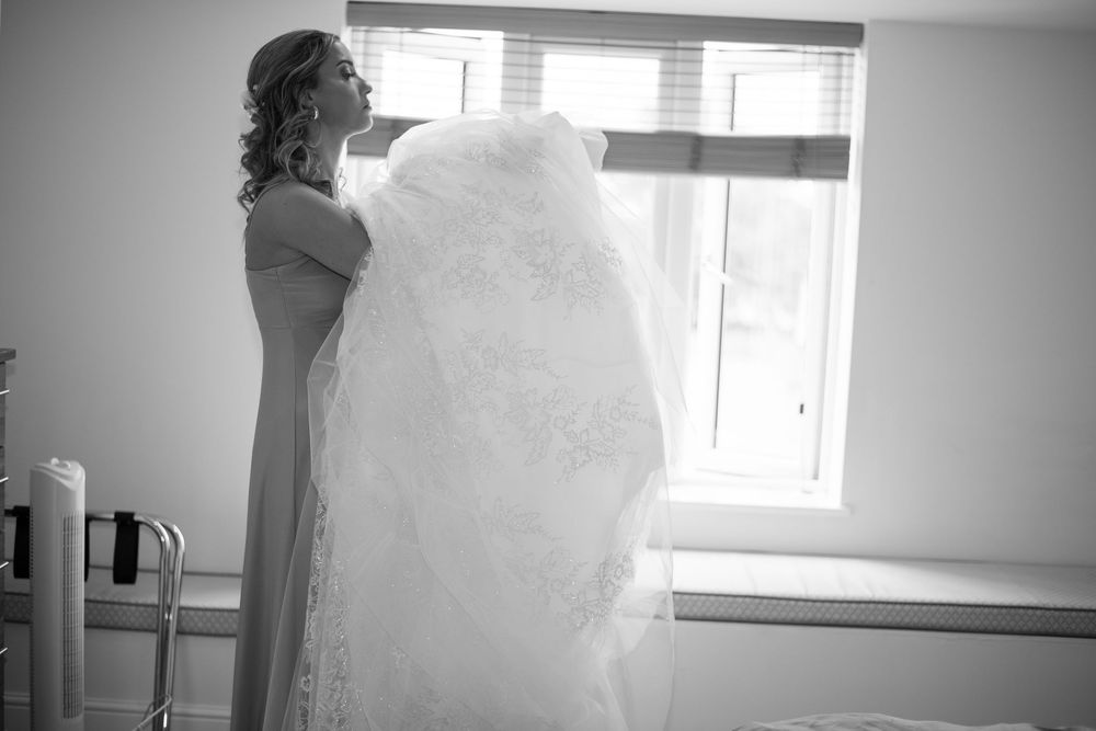 A white wedding dress being held up against natural window light in a bridal preparation room.