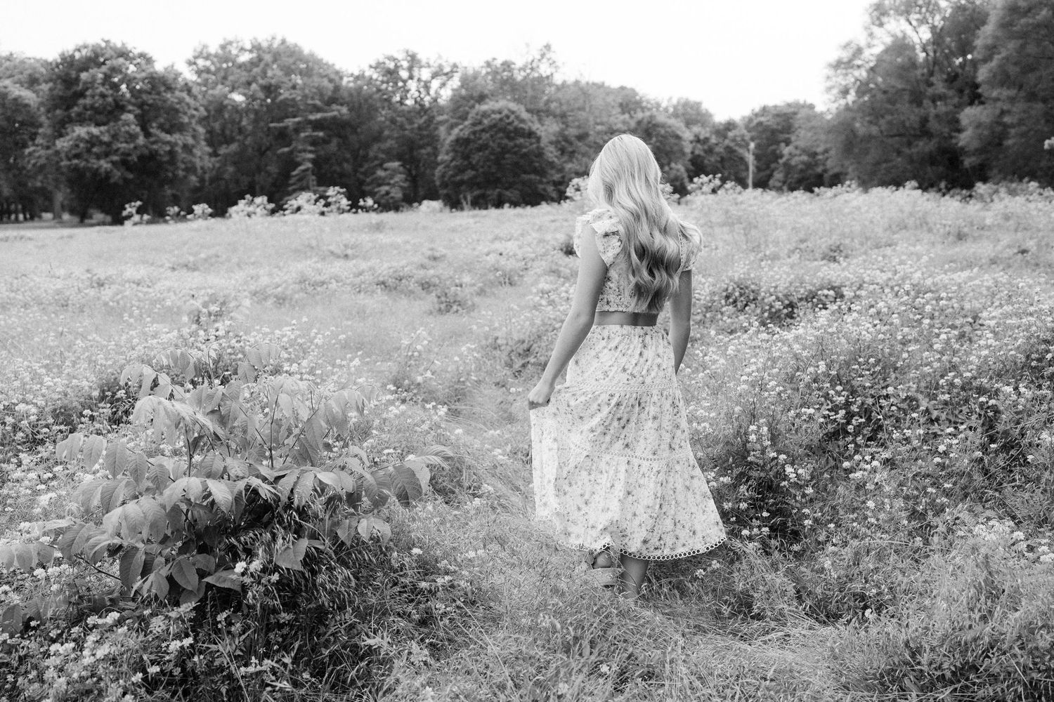 Woman in a flowing white dress walks through a sun-drenched wildflower meadow on a summer afternoon.