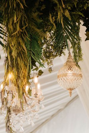 Elegant crystal chandeliers hang beneath lush cascading greenery and palm fronds at a luxury event venue.