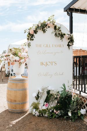 Elegant wedding welcome sign with gold lettering and floral decorations next to a rustic wine barrel at an outdoor venue.