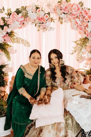 Two people sit together under a pink floral arch wearing elegant traditional South Asian attire.