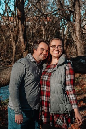 Couple wearing winter casual attire smiling together in a wooded outdoor setting during autumn.