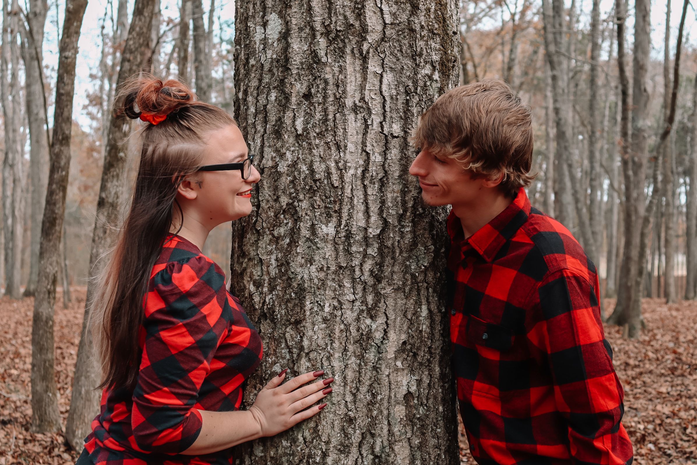 Couple wearing matching red buffalo plaid flannel shirts embracing in autumn forest setting.
