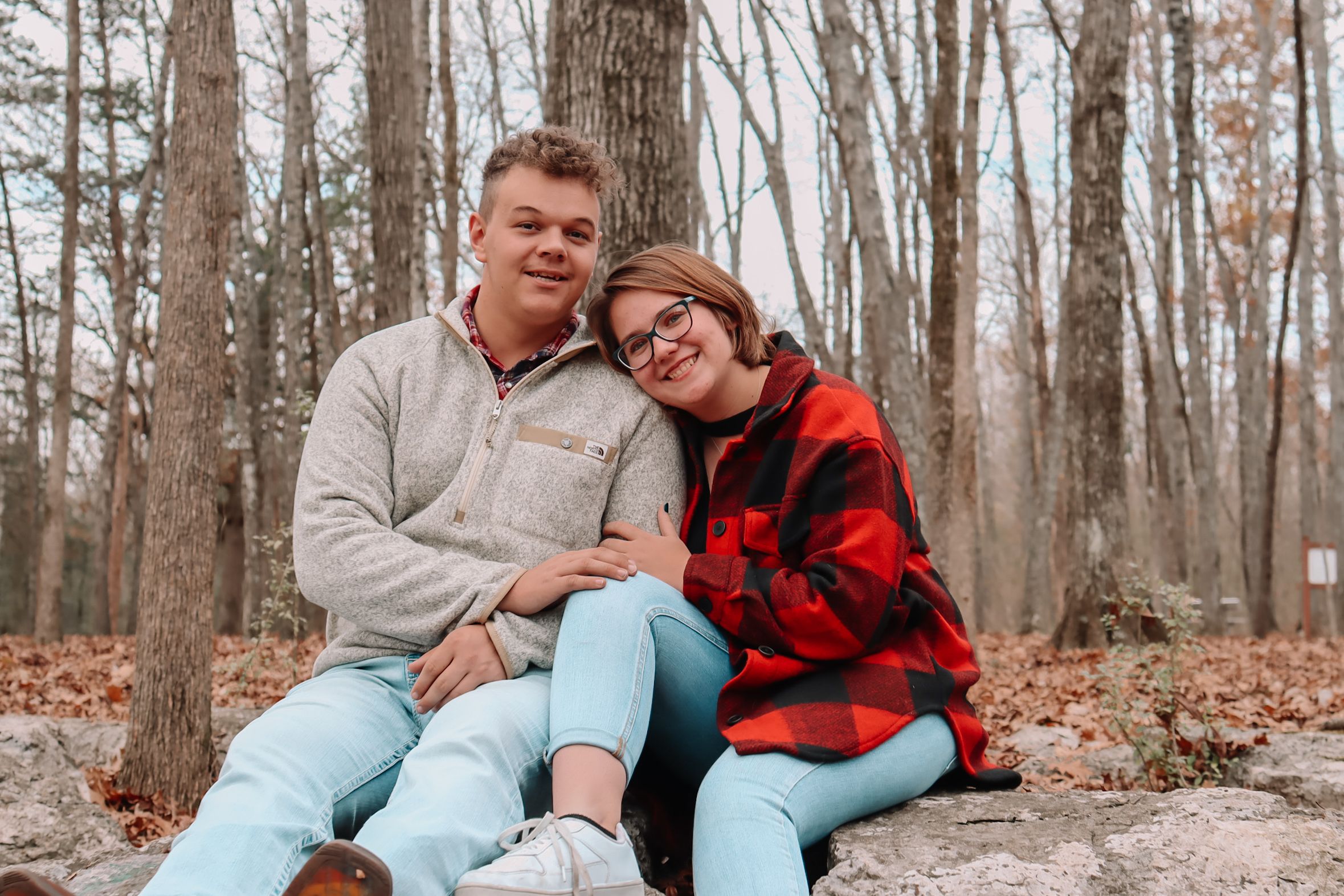 Couple in casual wear and red plaid sharing tender moments on forest floor with fallen leaves.