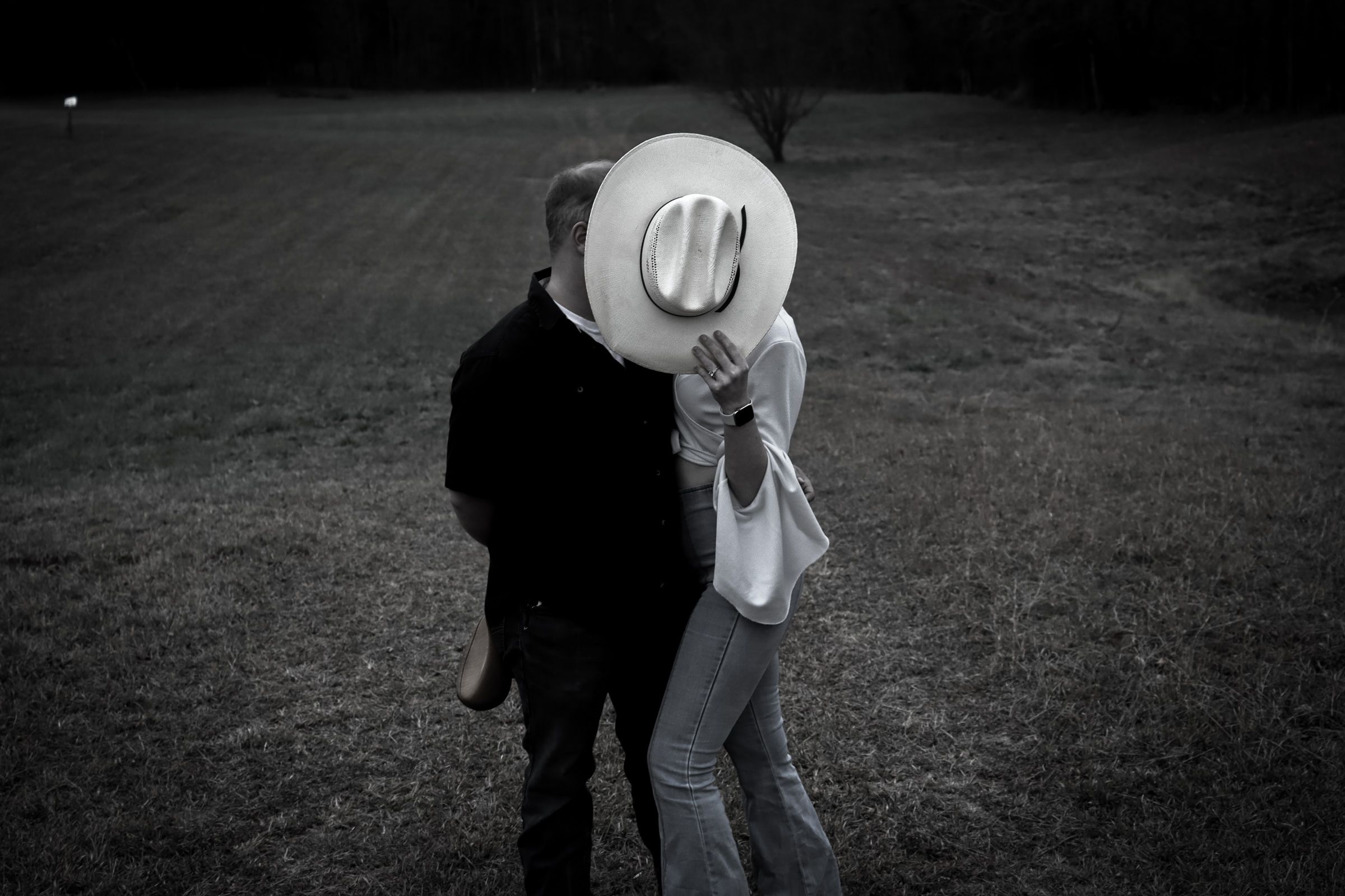 A couple shares an intimate moment in black and white photograph while person wearing cowboy hat covers their faces.