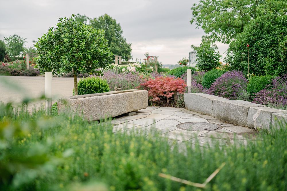 Steinmauer in einem üppigen Garten mit blühenden Pflanzen und Bäumen vor einem bewölkten Himmel.