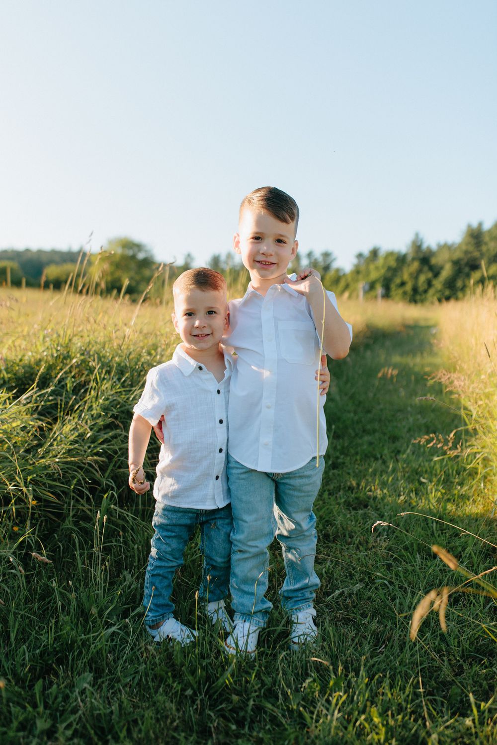 Two young children in white shirts and jeans stand together on a grassy path during golden hour.
