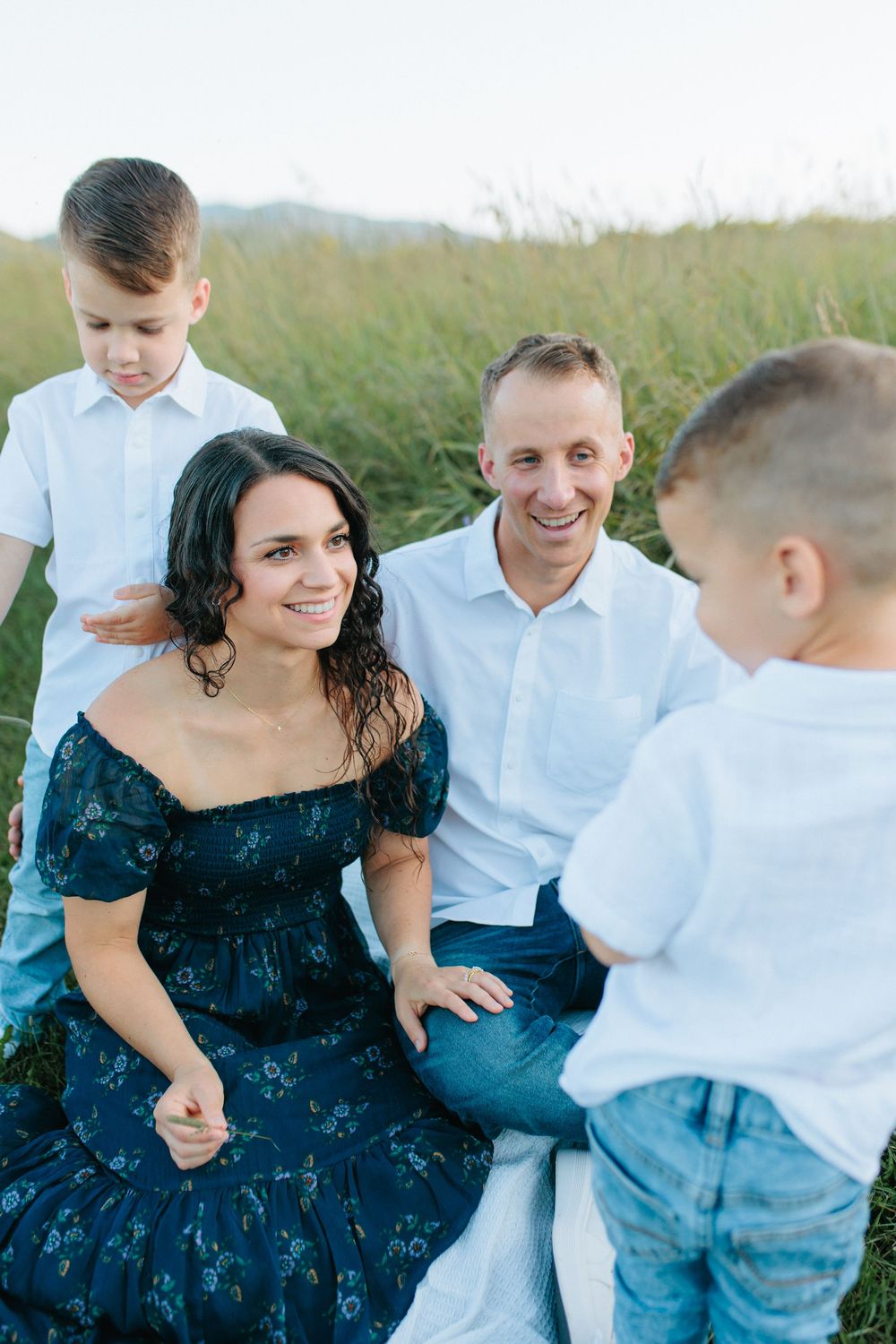 A woman in a dark blue dress sits outdoors in a field while interacting with someone in a white shirt.