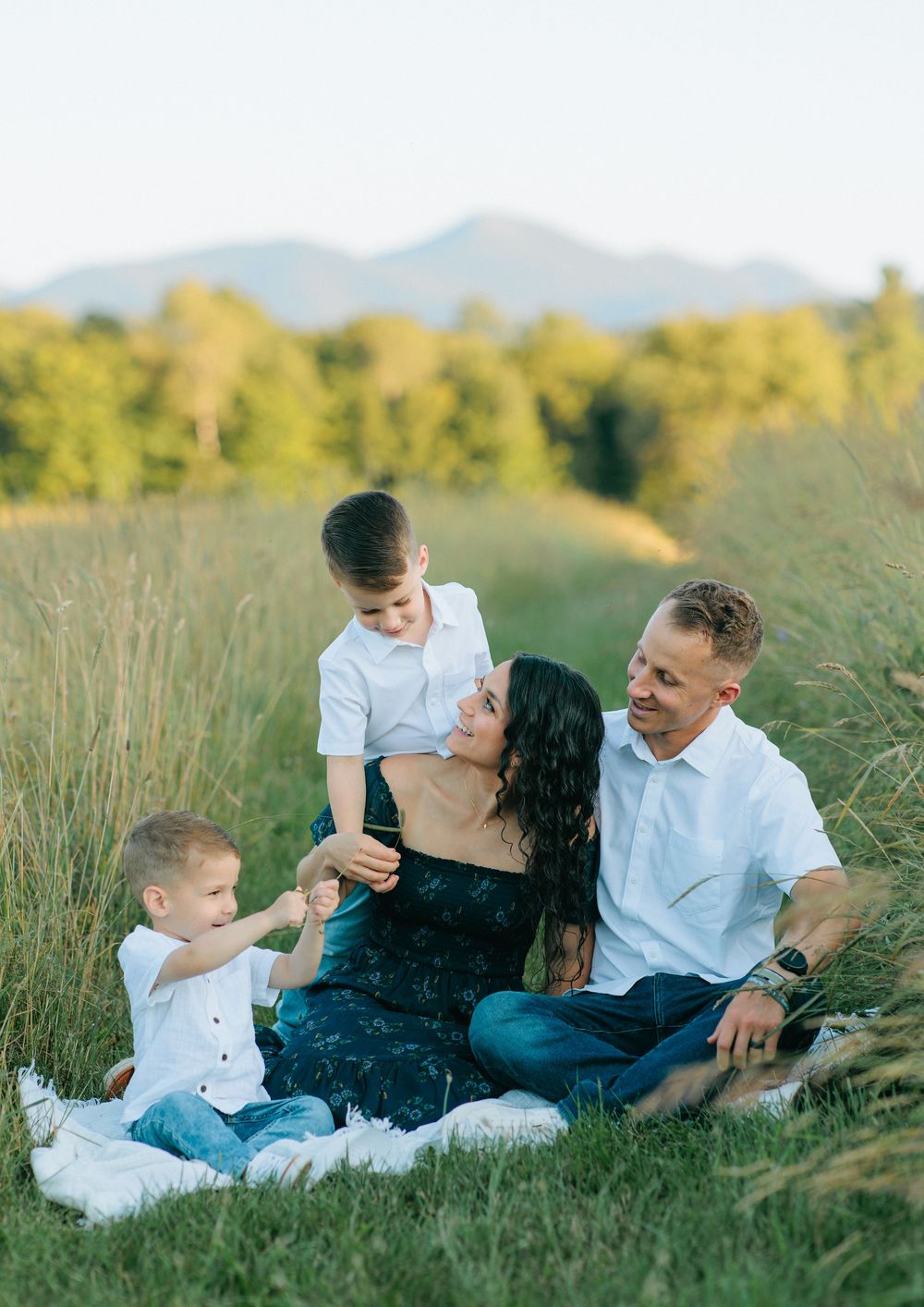 Family of four sits together on a blanket in a sunlit meadow with mountains and trees in the background.