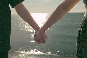 Two people hold hands while walking along a tranquil beach at sunset with ocean waves in the background.
