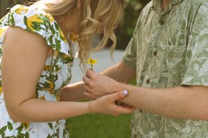 Close-up of two people holding hands outdoors, with a floral dress and green shirt visible in summertime.