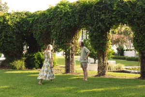 Two people enjoying a romantic moment under a leafy green garden arch in a sunny outdoor setting.