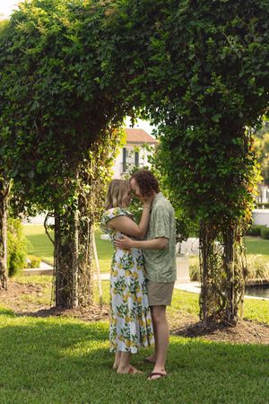 Two people sharing a romantic moment under a lush green garden archway on a sunny afternoon.