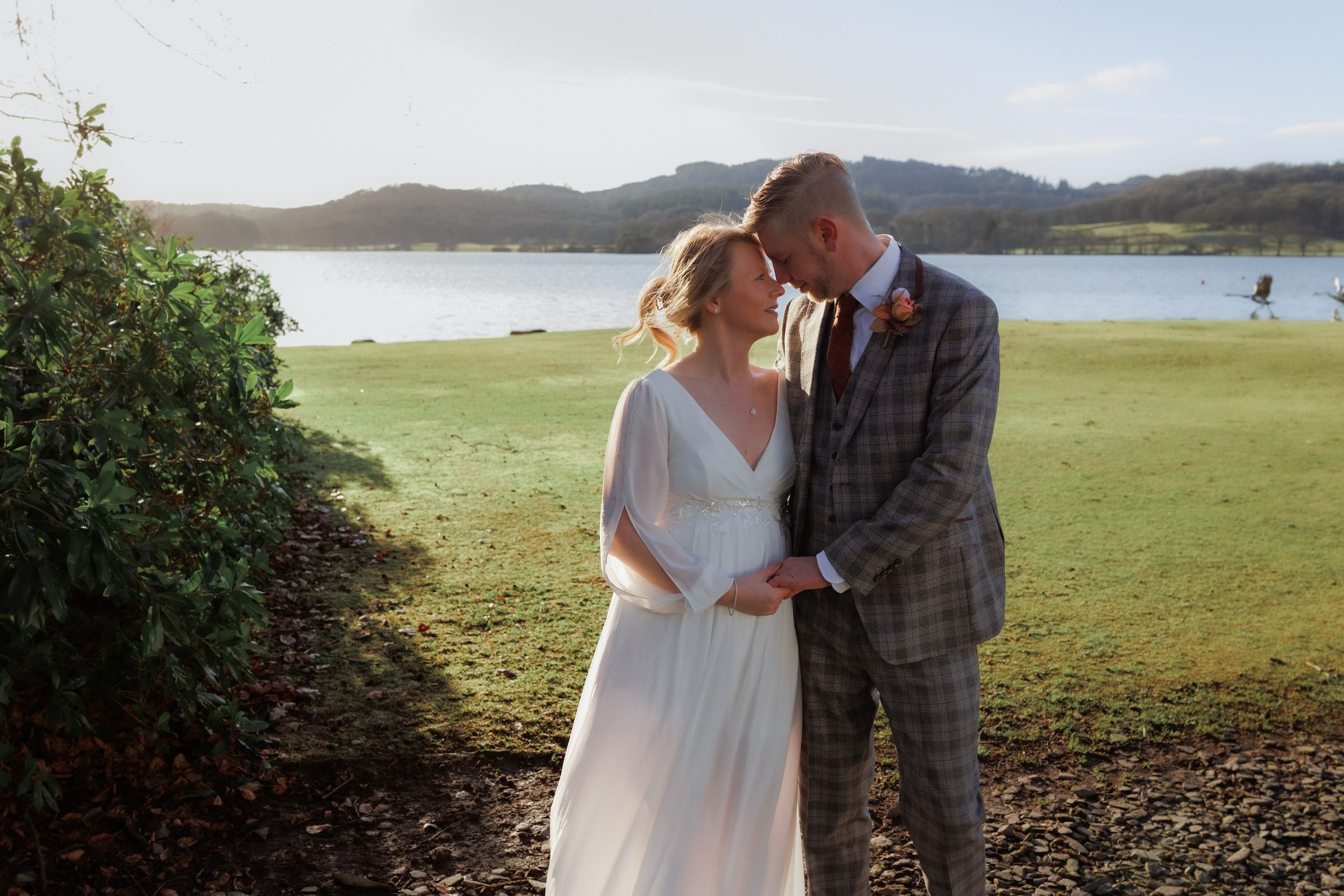 A couple shares a romantic kiss on a mountain trail overlooking a scenic lake and lush landscape during golden hour.