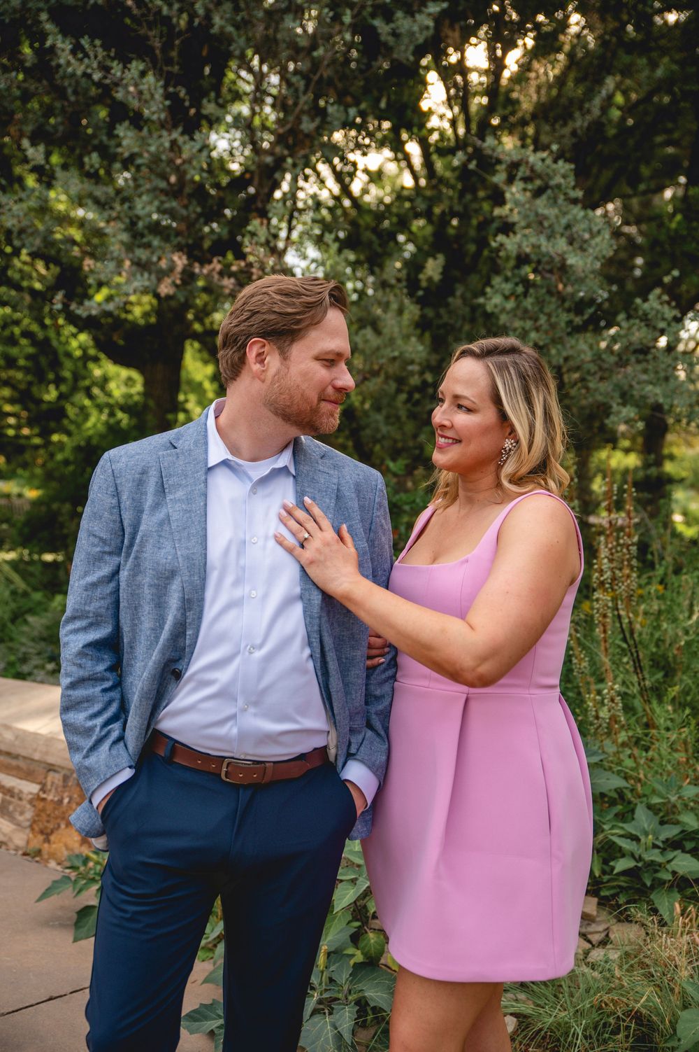 A couple embraces outdoors in a garden setting wearing coordinated formal attire with blue suit and pink top.