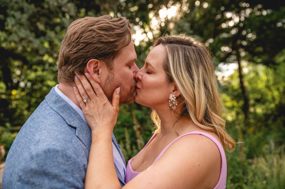 A couple shares tender kisses in an outdoor setting with lush greenery and natural sunlight in the background.