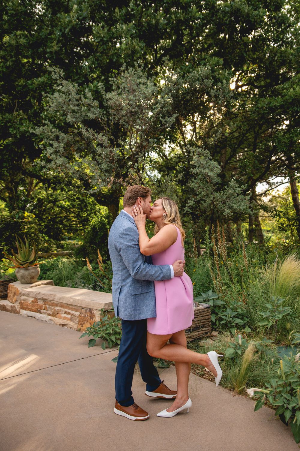 A couple sharing a romantic moment in a garden path during sunset, surrounded by greenery and stone walls.